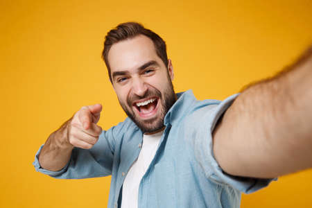 Close up of young man in casual blue shirt posing isolated on yellow orange background. People lifestyle concept. Mock up copy space. Doing selfie shot on mobile phone pointing index finger on camera.の写真素材