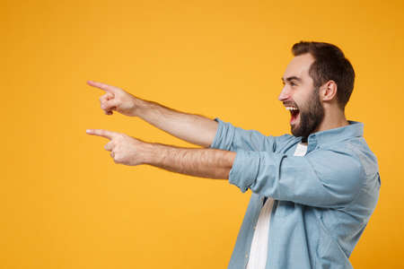 Side view of funny excited young bearded man in casual blue shirt posing isolated on yellow orange background in studio. People lifestyle concept. Mock up copy space. Pointing index fingers aside.の写真素材