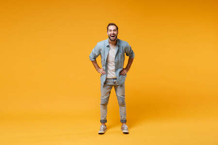 Laughing young bearded man in casual blue shirt posing isolated on yellow orange background, studio portrait. People emotions lifestyle concept. Mock up copy space. Standing with arms akimbo on waist.の写真素材