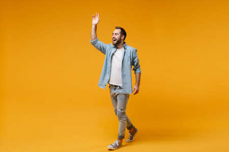 Cheerful young bearded man in casual blue shirt posing isolated on yellow orange background in studio. People lifestyle concept. Mock up copy space. Waving and greeting with hand as notices someone.の写真素材