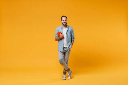 Smiling young student man in casual blue shirt posing isolated on yellow orange wall background studio portrait. People sincere emotions lifestyle concept. Mock up copy space. Holding red notebook.の写真素材
