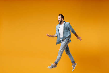 Cheerful young bearded man in casual blue shirt posing isolated on yellow orange wall background,...
