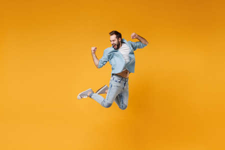 Happy young bearded man in casual blue shirt posing isolated on yellow orange background, studio portrait. People sincere emotions lifestyle concept. Mock up copy space. Jumping doing winner gesture.の写真素材