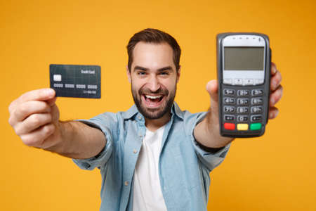 Cheerful young man in blue shirt posing isolated on yellow orange background. People lifestyle concept. Mock up copy space. Hold wireless bank payment terminal to process acquire credit card payments.の写真素材