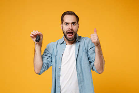 Shocked young man in casual blue shirt posing isolated on yellow orange background, studio portrait. People emotions lifestyle concept. Mock up copy space. Holding in hand car keys, showing thumb up.の写真素材