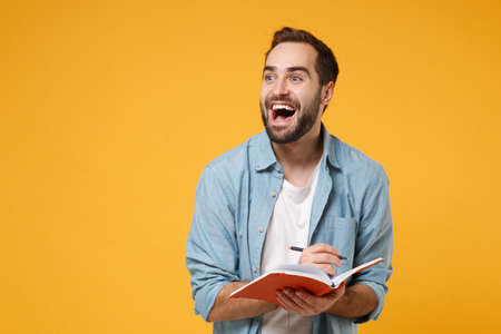 Excited young student man in casual blue shirt posing isolated on yellow orange wall background, studio portrait. People lifestyle concept. Mock up copy space. Writing note in notebook, looking aside.の写真素材