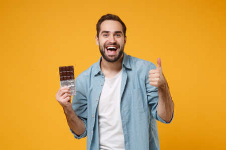 Cheerful young man in casual blue shirt posing isolated on yellow orange background studio portrait. People sincere emotions lifestyle concept. Mock up copy space. Hold chocolate bar showing thumb up.の写真素材