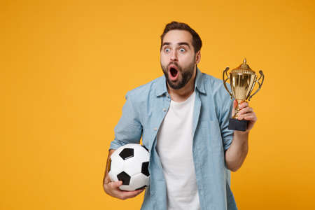 Shocked young man in casual blue shirt posing isolated on yellow orange wall background studio portrait. People lifestyle concept. Mock up copy space. Hold soccer ball football cup keeping mouth open.の写真素材