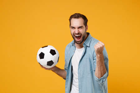 Joyful young man in blue shirt posing isolated on yellow orange wall background. People sincere emotions lifestyle concept. Mock up copy space. Holding soccer ball, doing winner gesture, screaming.の写真素材