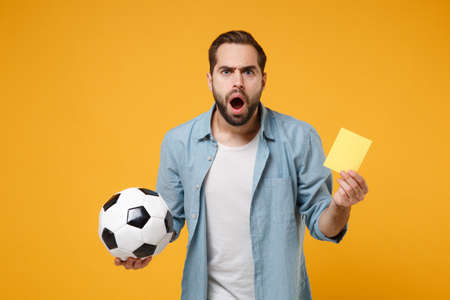 Shocked young man in casual blue shirt posing isolated on yellow orange wall background. People lifestyle concept. Mock up copy space. Hold soccer ball, yellow card, propose player retire from field.の写真素材