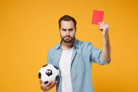 Dissatisfied young man in casual blue shirt posing isolated on yellow orange wall background. People lifestyle concept. Mock up copy space. Hold soccer ball, red card propose player retire from field.の写真素材