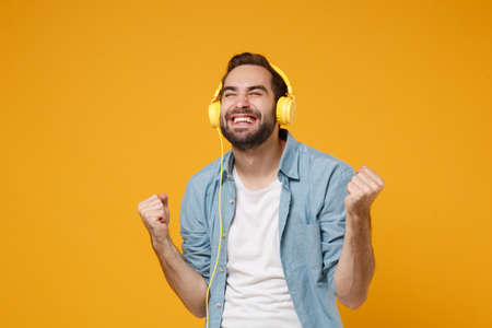 Happy young man in casual blue shirt posing isolated on yellow orange background, studio portrait. People lifestyle concept. Mock up copy space. Listen music with headphones, doing winner gesture.の写真素材