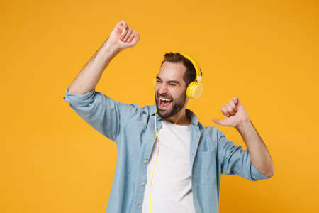 Cheerful young man in casual blue shirt posing isolated on yellow orange background, studio portrait. People lifestyle concept. Mock up copy space. Listen music with headphones, dancing, rising hands.の写真素材