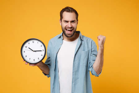 Joyful young man in casual blue shirt posing isolated on yellow orange background, studio portrait. People sincere emotions lifestyle concept. Mock up copy space. Holding clock, doing winner gesture.の写真素材