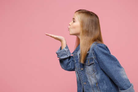 Side view of little blonde kid girl 12-13 years old in denim jacket isolated on pastel pink background children studio portrait. Childhood lifestyle concept. Mock up copy space. Blowing send air kiss.の写真素材
