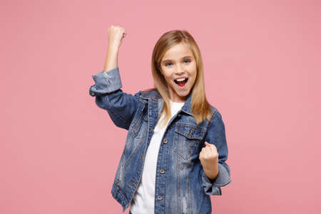 Happy little blonde kid girl 12-13 years old in denim jacket isolated on pastel pink background children studio portrait. Childhood lifestyle concept. Mock up copy space. Clenching fists like winner.の写真素材