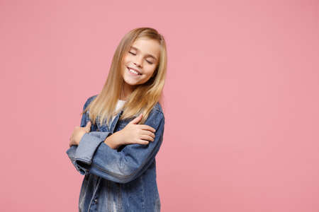 Smiling little blonde kid girl 12-13 years old in denim jacket isolated on pastel pink background in studio. Childhood lifestyle concept. Mock up copy space. Keeping eyes closed holding hands crossed.の写真素材
