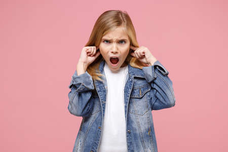 Shocked little kid girl 12-13 years old in denim jacket isolated on pastel pink background in studio. Childhood lifestyle concept. Mock up copy space. Keeping mouth open, covering ears with fingers.の写真素材