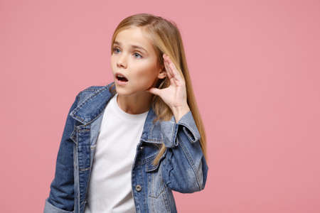 Curious little blonde kid girl 12-13 years old in denim jacket isolated on pastel pink background children portrait. Childhood lifestyle concept. Mock up copy space Try to hear you with hand near ear.の写真素材