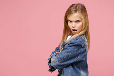 Side view of shocked little blonde kid girl 12-13 years old in denim jacket isolated on pastel pink background children portrait. Childhood lifestyle concept. Mock up copy space. Hold hands crossed.の写真素材