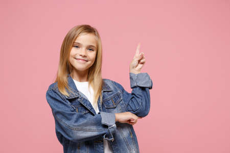 Smiling little kid girl 12-13 years old in denim jacket isolated on pastel pink background children studio portrait. Childhood lifestyle concept. Mock up copy space. Pointing index finger aside up.の写真素材
