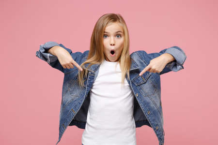 Shocked little blonde kid girl 12-13 years old in denim jacket isolated on pastel pink wall background. Childhood lifestyle concept. Mock up copy space. Pointing index fingers down keeping mouth open.の写真素材