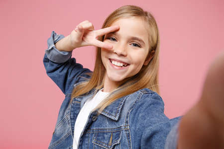 Close up of little kid girl 12-13 years old in denim jacket isolated on pastel pink wall background. Childhood lifestyle concept. Mock up copy space. Doing selfie on mobile phone showing victory sign.の写真素材