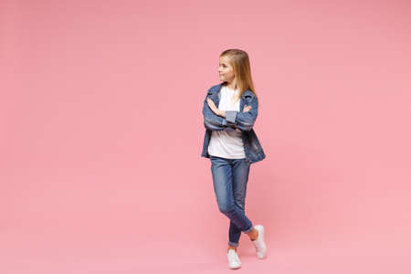 Smiling little blonde kid girl 12-13 years old in denim jacket isolated on pastel pink background children portrait. Childhood lifestyle concept. Mock up copy space. Hold hands crossed, looking aside.の写真素材