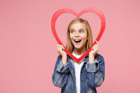 Cheerful little kid girl 12-13 years old in denim jacket isolated on pastel pink background children portrait. Childhood lifestyle concept. Mock up copy space. Hold red big wooden heart looking aside.の写真素材