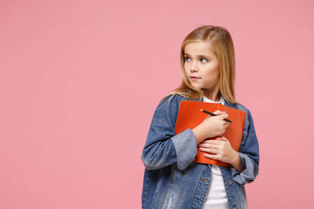 Little blonde kid girl 12-13 years old in denim jacket isolated on pastel pink background children studio portrait. Childhood lifestyle concept. Mock up copy space. Hold notebook pencil looking aside.の写真素材