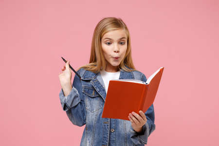 Amazed little blonde kid girl 12-13 years old in denim jacket isolated on pastel pink background children studio portrait. Childhood lifestyle concept. Mock up copy space. Holding notebook, pencil.の写真素材
