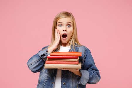 Shocked little kid girl 12-13 years old in denim jacket isolated on pastel pink background children portrait. Childhood lifestyle concept. Mock up copy space. Hold books, notebooks, put hand on cheek.の写真素材