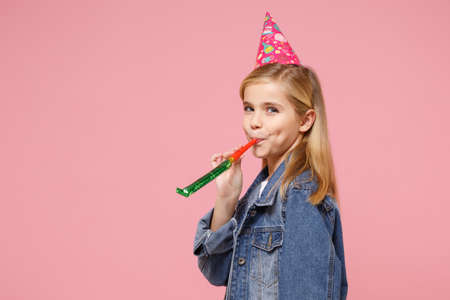 Side view of pretty little kid girl 12-13 years old in denim jacket, birthday hat isolated on pastel pink background in studio. Childhood lifestyle concept. Mock up copy space. Blowing on party pipe.の写真素材
