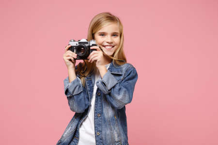 Smiling little blonde kid girl 12-13 years old in denim jacket posing isolated on pastel pink background in studio. Childhood lifestyle concept. Mock up copy space. Hold retro vintage photo camera.の写真素材