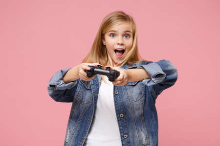 Excited little blonde kid girl 12-13 years old in denim jacket posing isolated on pastel pink background children portrait. Childhood lifestyle concept. Mock up copy space. Play game with joystick.の写真素材