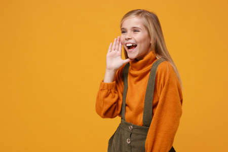 Laughing little kid girl 12-13 years old in turtleneck, jumpsuit isolated on orange yellow background. Childhood lifestyle concept. Mock up copy space. Whispering secret behind her hand, sharing news.の写真素材
