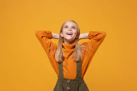 Pensive little blonde kid girl 12-13 years old in turtleneck, jumpsuit isolated on orange yellow wall background. Childhood lifestyle concept. Mock up copy space. Looking up with hands behind head.の写真素材