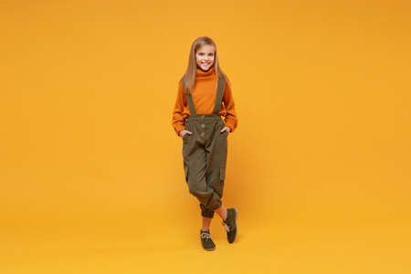 Smiling little kid girl 12-13 years old in turtleneck jumpsuit isolated on orange yellow background children studio portrait. Childhood lifestyle concept. Mock up copy space. Holding hands in pockets.の写真素材