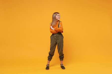 Funny little kid girl 12-13 years old in turtleneck jumpsuit isolated on orange yellow wall background in studio. Childhood lifestyle concept. Mock up copy space. Holding hands crossed, looking aside.の写真素材