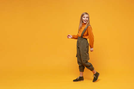 Laughing little blonde kid girl 12-13 years old in turtleneck jumpsuit isolated on orange yellow background children studio portrait. Childhood lifestyle concept. Mock up copy space. Looking camera.の写真素材