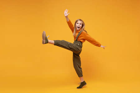 Cheerful little blonde kid girl 12-13 years old in turtleneck, jumpsuit isolated on orange yellow background. Childhood lifestyle concept. Mock up copy space. Dancing, rising leg, spreading hands.の写真素材