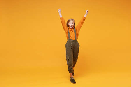 Joyful little blonde kid girl 12-13 years old in turtleneck jumpsuit isolated on orange yellow background in studio. Childhood lifestyle concept. Mock up copy space. Rising hands doing winner gesture.の写真素材