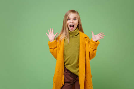 Excited little blonde kid girl 12-13 years old in yellow coat posing isolated on pastel green wall background. Childhood lifestyle concept. Mock up copy space. Keeping mouth open, spreading hands.の写真素材