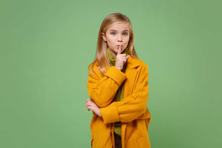 Secret little kid girl 12-13 years old in yellow coat posing isolated on pastel green background. Childhood lifestyle concept. Mock up copy space. Say hush be quiet with finger on lips shhh gesture.の写真素材