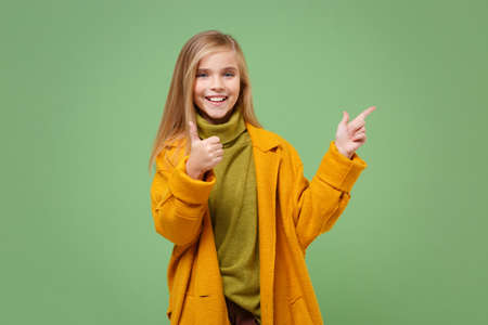 Funny little blonde kid girl 12-13 years old in yellow coat posing isolated on pastel green background. Childhood lifestyle concept. Mock up copy space. Pointing index finger aside, showing thumb up.の写真素材