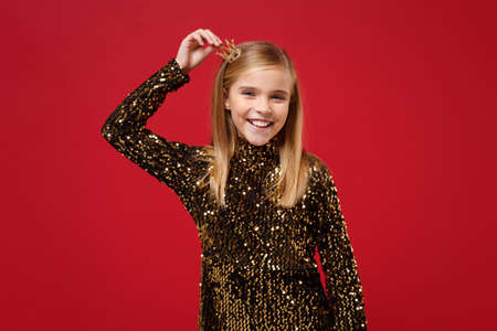 Cheerful little blonde kid girl 12-13 years old in glitter party outfit, crown isolated on red background children studio portrait. Childhood lifestyle concept. Mock up copy space. Put hand on head.の写真素材