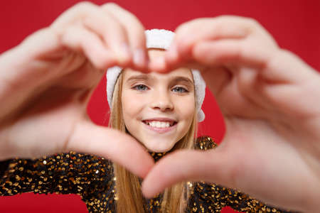 Smiling little kid girl 12-13 years old in glitter party outfit Santa hat isolated on red background. New Year 2020 celebration holiday concept. Mock up copy space. Showing shape heart with hands.の写真素材