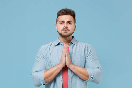 Pleading young bearded guy 20s in casual shirt posing isolated on pastel blue wall background studio portrait. People emotions, lifestyle concept. Mock up copy space. Holding hands folded in prayer.の写真素材