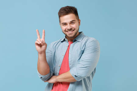 Smiling funny young bearded guy 20s in casual shirt posing isolated on pastel blue wall background studio portrait. People sincere emotions lifestyle concept. Mock up copy space. Showing victory sign.の写真素材