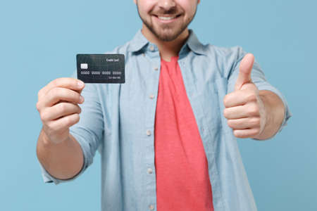Cropped image of smiling young bearded guy 20s in casual shirt posing isolated on pastel blue background studio. People lifestyle concept. Mock up copy space. Hold credit bank card, showing thumb up.の写真素材